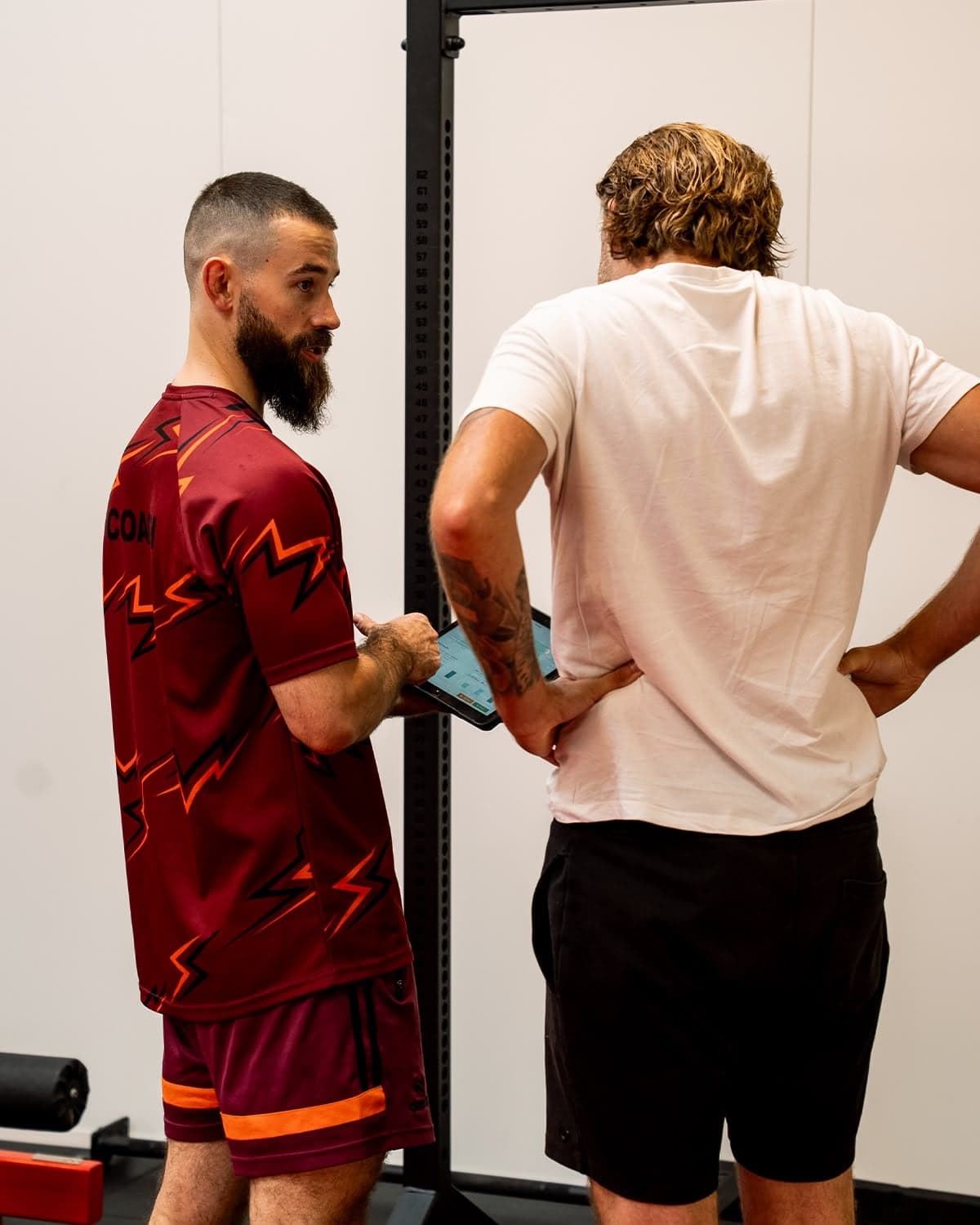 A Magni Fitness coach in branded kit recording assessment numbers on a tablet with a member, beside the height scale on the rig.
