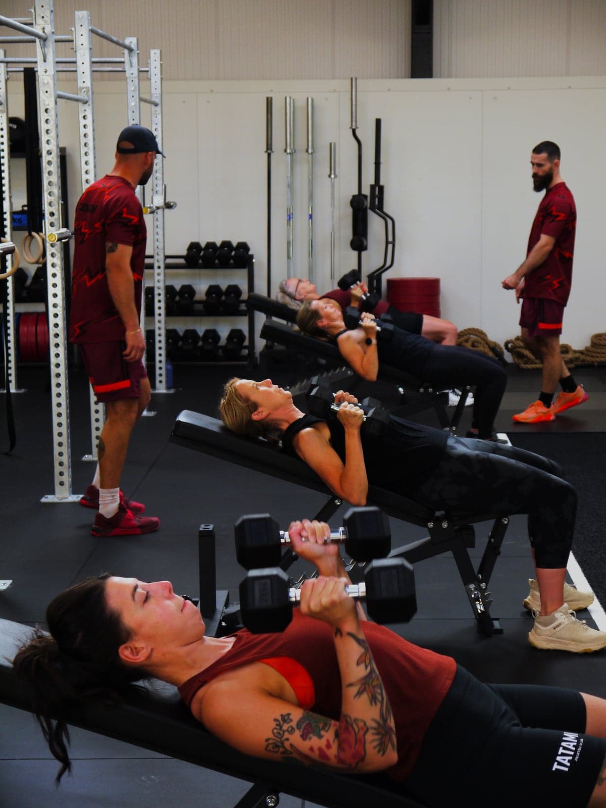 Members pressing dumbbells on a row of benches during a group personal training session, with two coaches in branded kit watching their form.