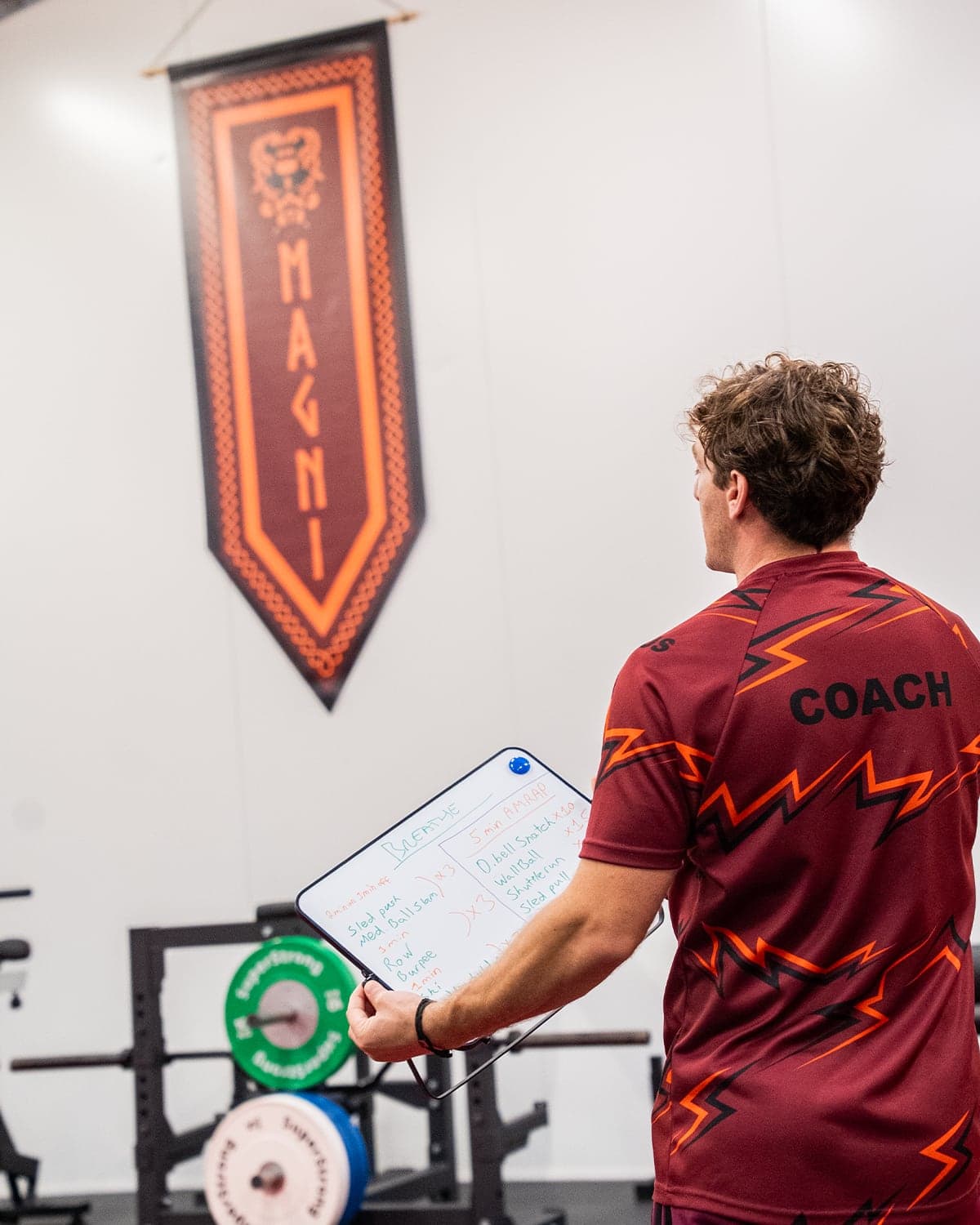 A Magni Fitness coach in branded kit holding a whiteboard with the Breathe session written out, beneath the MAGNI banner on the gym wall.