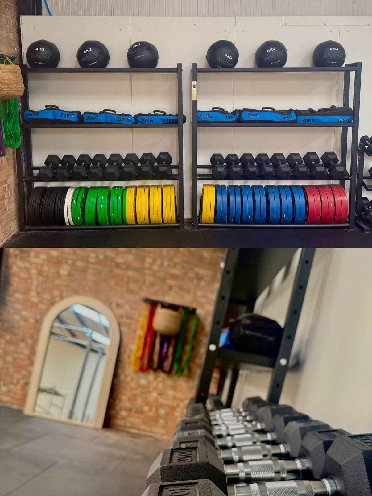 Weight storage racks loaded with medicine balls, sandbags, dumbbells, and colour bumper plates, above a row of dumbbells leading to the arched mirror and resistance bands.