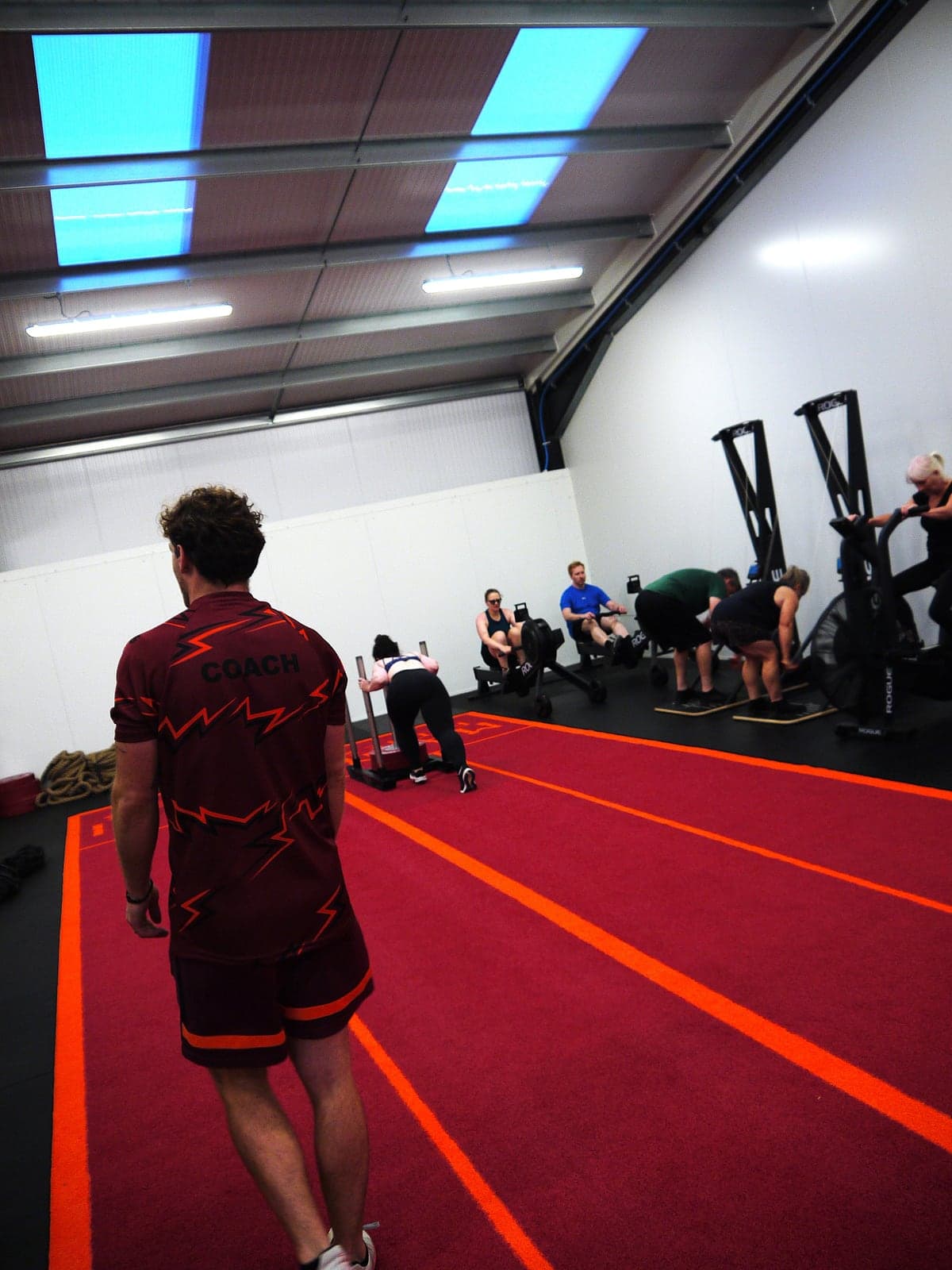 A coach watching a conditioning session from the red sled track, with members working on the rowers, bikes and a loaded sled.