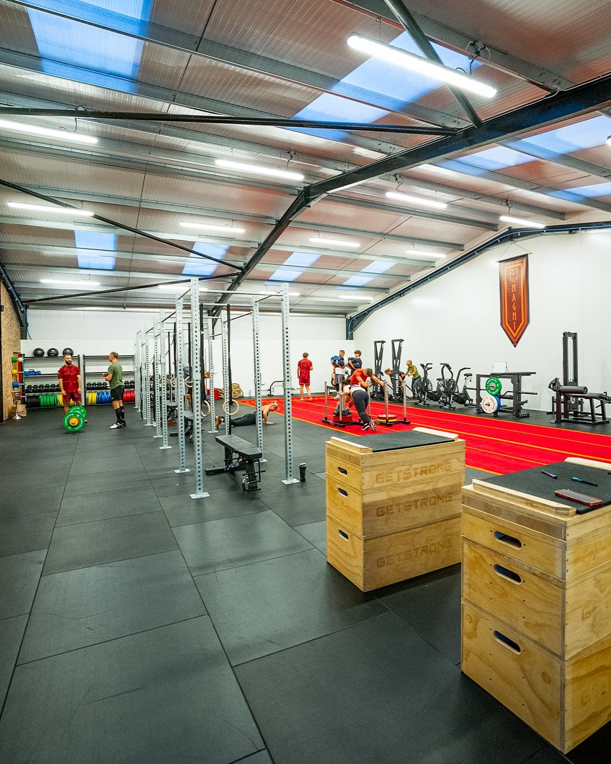 A wide view down the Magni Fitness gym floor: the run of lifting rigs, the red sled track with members training, and the MAGNI banner on the far wall.