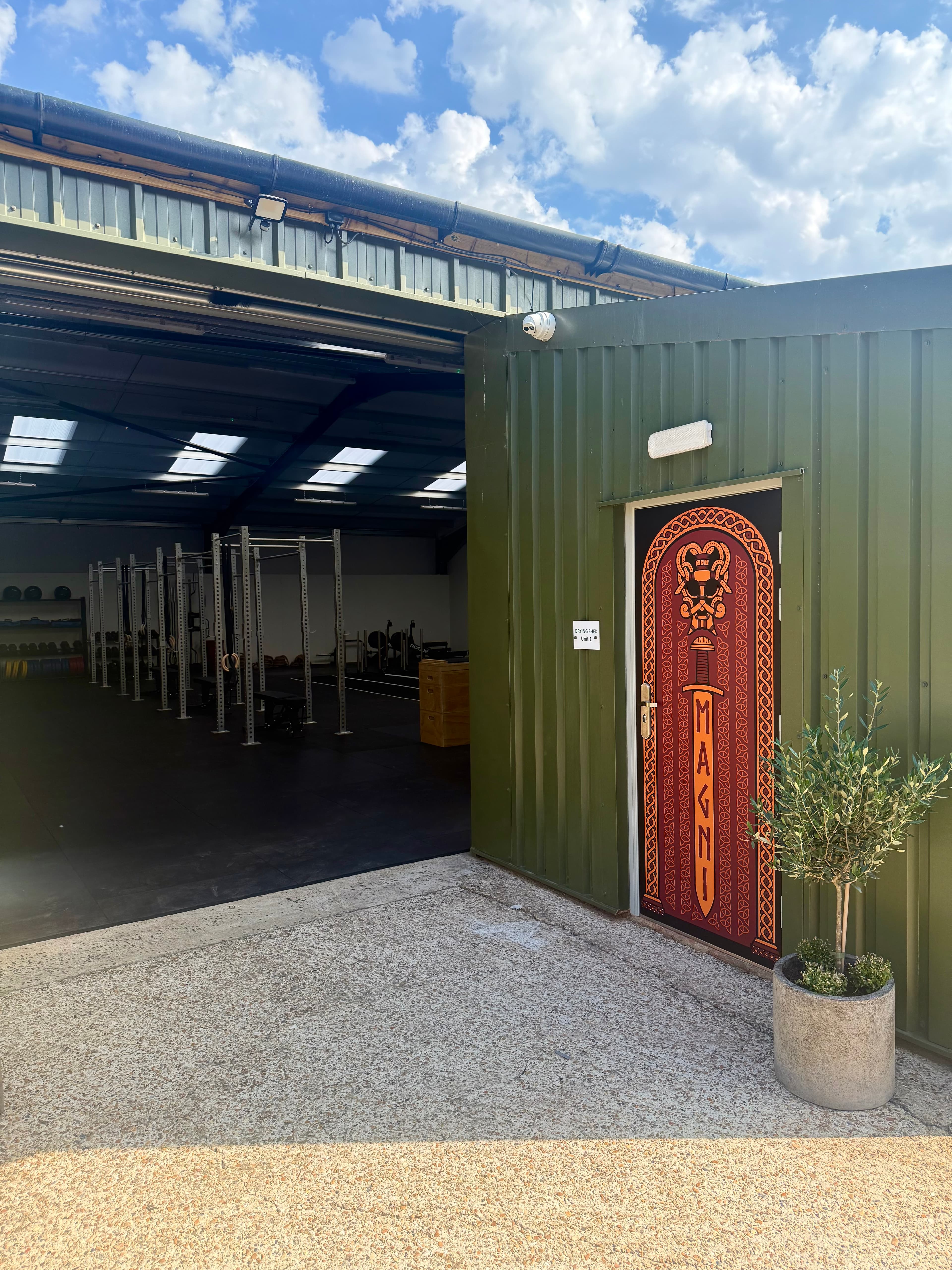 The green-clad exterior of the Magni Fitness unit, with the branded Nordic-style door reading MAGNI and the gym floor visible through the open bay door.