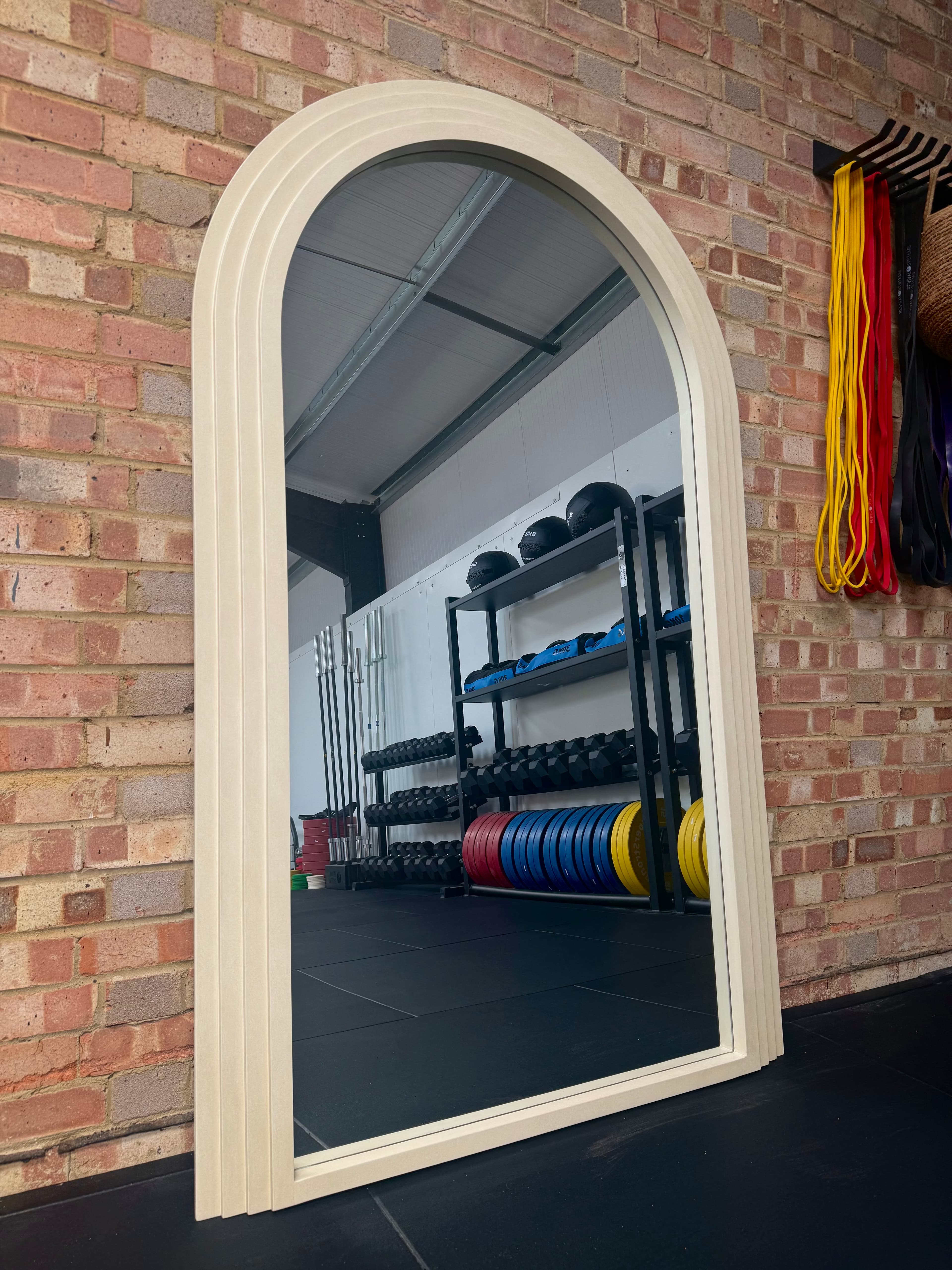 Arched cream-framed mirror against the brick wall, reflecting the dumbbell racks, medicine balls, and colour bumper plates.