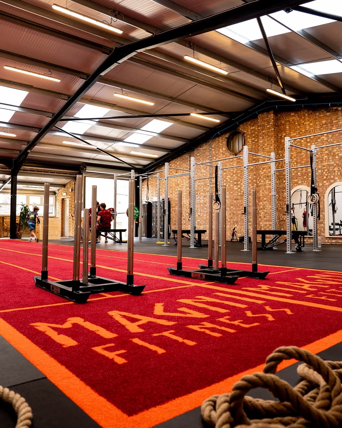 The red Magni Fitness sled track running the length of the gym floor, with loaded sleds on the turf and the lifting rigs and brick wall behind.