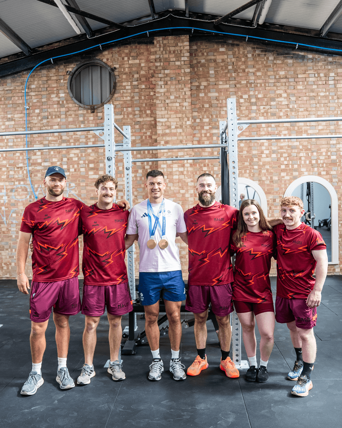 The Magni Fitness coaching team in branded kit, standing together with a guest wearing Team GB apparel and medals, on the gym floor.